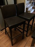 Two dark brown faux leather high top chairs with wooden legs, positioned side by side on wooden floor.
