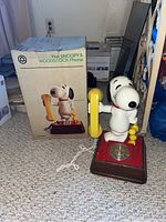 Photo shows the Snoopy and Woodstock phone with original box, Snoopy holding yellow handset, Woodstock by his side, red and wood grain base with rotary dial.