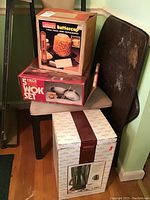 Overall lot photo showing boxed Montgomery Ward Buttercup popcorn maker on top, boxed Himark 5-piece wok set below, and boxed Gevalia coffee maker at bottom, all stacked on a chair near a wooden tray or board