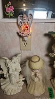 Royal Albert plug-in night light glowing with angel and floral design on wall outlet; below on counter two angel figurines, one white ceramic, one with blond hair.