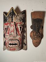 Photo of two antique tribal African hand carved masks side by side on a cream background, showing weathered paint and wood texture.