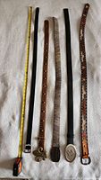 Overview of all five leather belts laid out side-by-side, showing various buckles and lengths against a white towel background with measuring tape beside them.