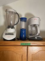 Kitchen appliances on wooden surface: left Sunbeam mixer, middle blue Hamilton Beach personal blender, right Sunbeam coffee maker with glass carafe