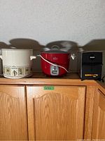 Photo of three kitchen appliances on wooden cabinet: white Rival Crock Pot slow cooker with leaf design, red rice cooker, and black Silex two-slice toaster.