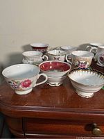 View of eight tea cups with various floral, gold, and geometric decorative patterns arranged on wood surface.