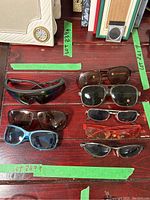 Top-down view of eight assorted sunglasses of different styles and lens colors laid out on wooden surface with green tape labeling.