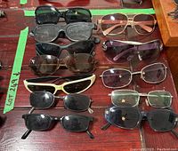 Top-down view of lot showing 12 assorted sunglasses varied in frame size, color, and lens tint. All items laid flat on reddish wood surface.