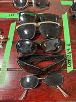 Top down view showing all 5 pairs of assorted black frame sunglasses laid flat on a dark wood surface with green tape labels.