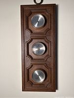 Full front view of brown wooden weather station with three silver round dials for measuring weather conditions, set vertically in rectangular carved wooden panel.