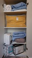 White and pastel blue folded linens on shelves with mustard yellow corduroy pillow in middle shelf, white packaged items and light blue fleece blanket below.