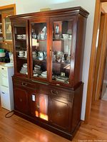 Front and angle views of dark wood buffet and hutch cabinet with glass doors on upper section displaying dishes and collectibles, and solid wood doors on lower section.