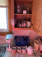 Photo of white Betty Crocker drip coffee maker on kitchen counter next to multiple mugs and teapot, with glass salad bowls and shakers inside wood kitchen cabinet