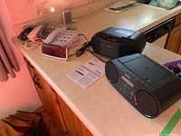 Overview photo showing a black Sony top-loading CD/radio, a black Sony boombox CD/radio, a red digital alarm clock, and two white telephones with large buttons on a countertop.