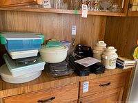 Photo showing various kitchen items on wooden shelves including plastic food storage containers, Pyrex bakeware, metal meat grinder, ceramic canisters, cookbooks and electric cutting tools.