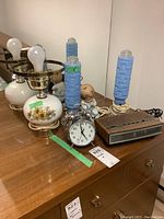 Photo of two vintage floral glass lamps with metal fixtures, blue textured glass lamps, ceramic praying child lamp, metal alarm clock and GE clock radio on wooden surface.