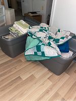 Two large plastic totes filled with assorted bed linens and a handmade quilt draped on top