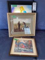 Three framed prints stacked: a 11x15 religious hologram print at top in dark brown frame, 8x9.5 farmers in field in gold frame, and 7x9 red house by river print in brown frame.