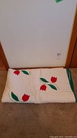 Folded white twin quilt with red tulips and green leaves against brown carpet and white wall, showing green trim along edge.