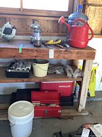 Wide view of utility tools on workbench including red watering can, blue water jug, battery lanterns, gardening tools, toolbox and cooler below.