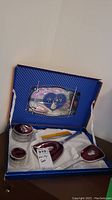 Photo showing open vanity set with two lidded glass dishes, brush, comb, and mirror on white satin lining inside original blue striped box. The mirror is mounted on the box lid with blue heart and floral design.