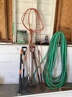 Yard tools including shovel, pitchfork, rake, garden hoe leaning against white wall. Coiled green garden hose, orange extension cord and bag of grass seed present.
