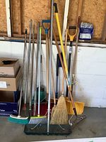 Photo showing various long-handled yard tools including brooms, rakes, and shovels arranged against a concrete wall, with boxes stacked to the side.