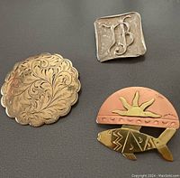 Three brooches displayed on black, showing gold filled engraved round brooch, sterling silver square JB monogram brooch and artisan copper & brass fish brooch.