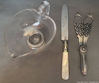 Three items displayed together: a clear glass sauce boat with a sterling silver base, a silver plate cake cutter with mother of pearl handle, and silver plate grape scissors with ornate filigree handles.