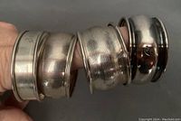 Four metal napkin rings shown stacked on fingers, showing varied designs and metal types.