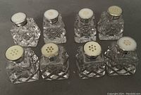 Eight clear cut crystal salt shakers with sterling silver and mother of pearl tops arranged in two rows, showing tops and crystal bodies.