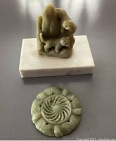 Green stone sculpture of two kittens or cubs on a marble base and a separate carved circular green stone disc laid on a flat surface.