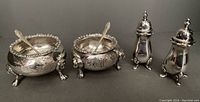 Two silver plate open salts with claw feet and matching floral engraving, two small matching salt spoons placed inside salts, and two small plate salt shakers with dome lids on right.