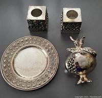 Four silver Judaica items displayed: two square candle holders with detailed Jerusalem panorama, silver pomegranate figurine, and round small dish with geometric border.