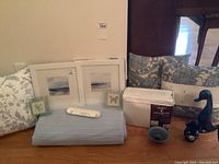 Wide shot of all items showing decorative pillows, sheet set, framed prints, cotton blanket, and blue ceramic bird with trivets.