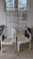 Two white resin chairs placed in front of two black metal wire trellises in a garage setting.