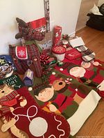 Wide view showing plaid reindeer figure, red felt cat stocking with yarn ball detail, fabric chair backings with elf and bear faces, gift bags including snowman and 'good cat', sticky bows, and greenery decorations on floor.