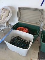 Two large plastic totes; one white filled with Christmas string lights, one green containing extension cords and flood lights; garage floor setting