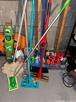 Wide view of various cleaning tools and supplies showing mops, broom, Swiffer box, and assorted spray bottles on basement floor with concrete wall