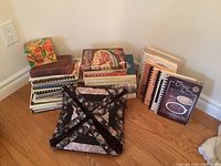 Wide shot of stacked cookbooks and accessories on the floor, includes recipe box, spiral-bound and hardcover cookbooks.