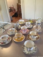 Photo of 12 fine bone china tea cups and saucers arranged on kitchen counter, varied floral and gold designs.