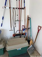 Long handled gardening and cleaning tools hanging and leaning against wall including rakes, shovels, broom, hoe, and an extendable pole broom alongside a large plastic storage tote on the floor.