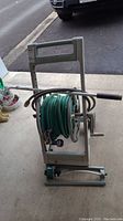 Front view of Ames HoseKing hose reel loaded with green garden hose and spray nozzle attached to the hose end. Shows hose reel handle, crank, and wheels.