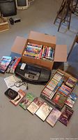 Box filled with VHS tapes, several cassette tapes laid out, a Panasonic black cassette radio, and a compact cassette player on a floor.