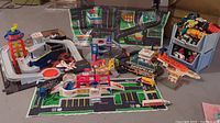 Wide view showing plastic road mats spread on the floor with various Matchbox play sets arranged including the airport, hospital, parking structure, and carwash. Several toy vehicles and small figures placed around the sets.