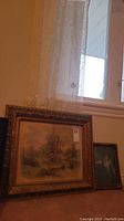 Two framed prints pictured leaning against the wall under a window with lace curtains. The larger print shows a farmhouse scene and the smaller one a woman with a rabbit.