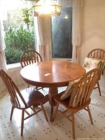 View of the round oak dining table with four Windsor chairs placed around it, each chair with cushions on seat backs and seating areas. The table has a smooth top with visible wood grain.