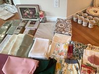 Multiple folded tablecloths in various colors including white, green, pink, floral patterns, laid out on floor with cloth napkins and aprons niche