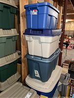 Stack of five Rubbermaid storage totes in basement setting: three stacked blue, white, and grey totes, with beige totes stacked next to them.