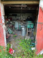 View into shed showing wooden shelves with various garden tools, metal trash can, and overgrowth at the entrance.