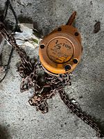 Top-down view of the orange metal chain hoist with attached rusty chain on concrete ground.